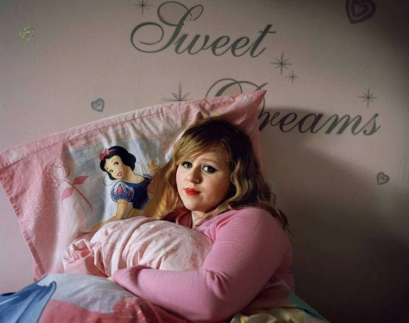 Young woman with long hair in bed with pink sheets and cartoon pillow. Pink wall has 'Sweet Dreams' text and heart decals.
