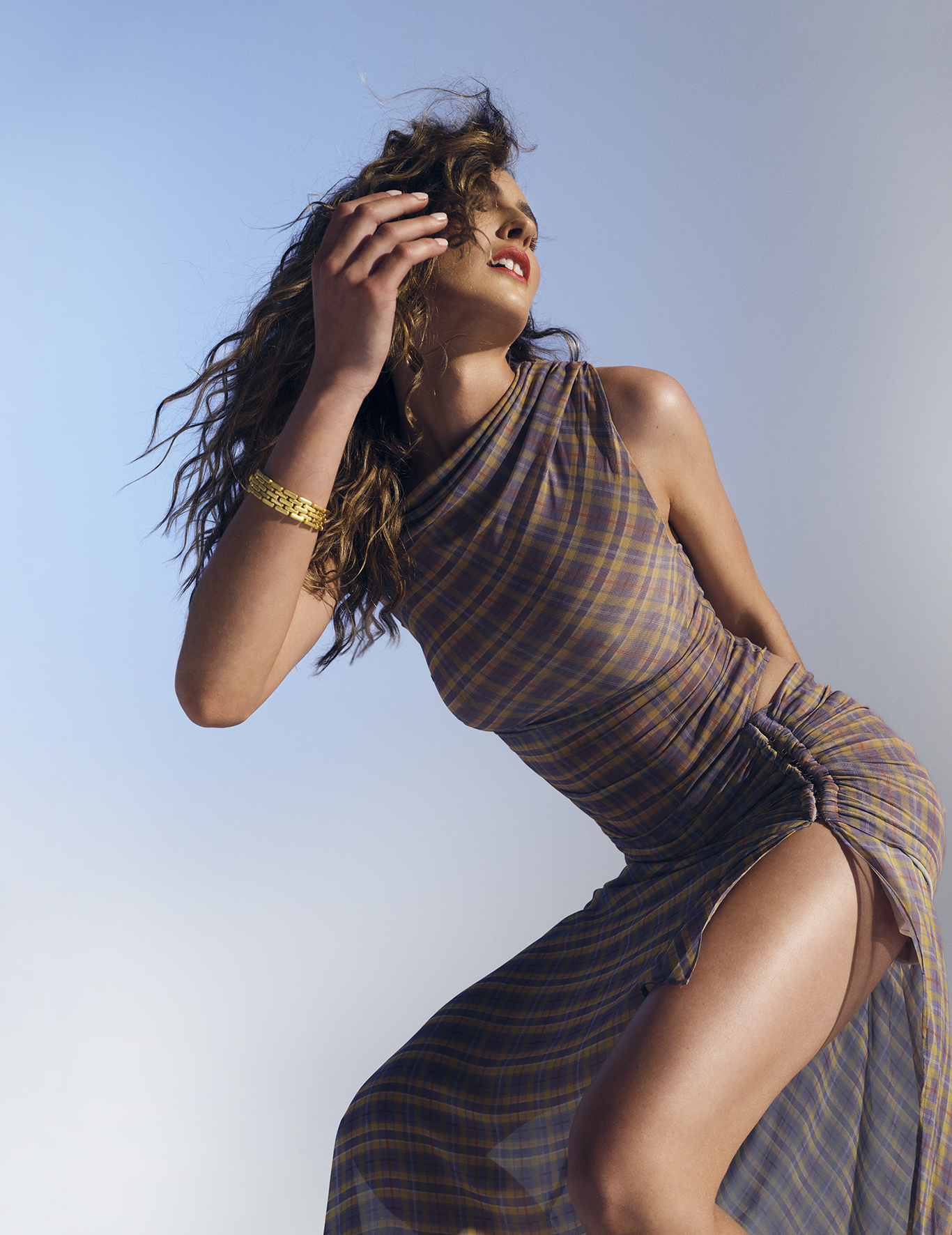 An adult woman with curly hair in a plaid dress poses against a blue-and-white background. The dress has a thigh slit.