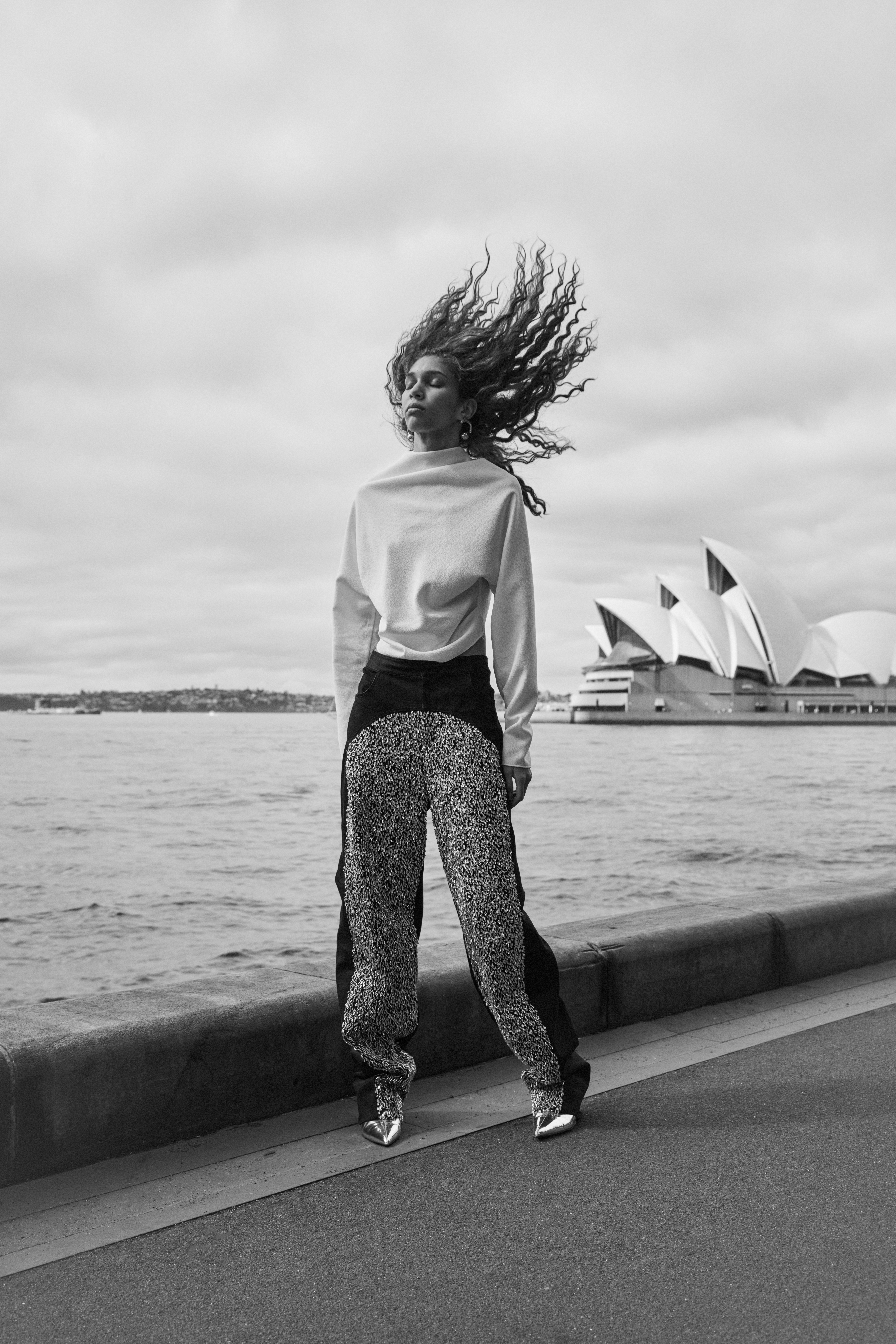 Black and white outdoor fashion shot of a woman with curly hair wearing a mock neck sweater and patterned pants, with the Sydney Opera House in the background.