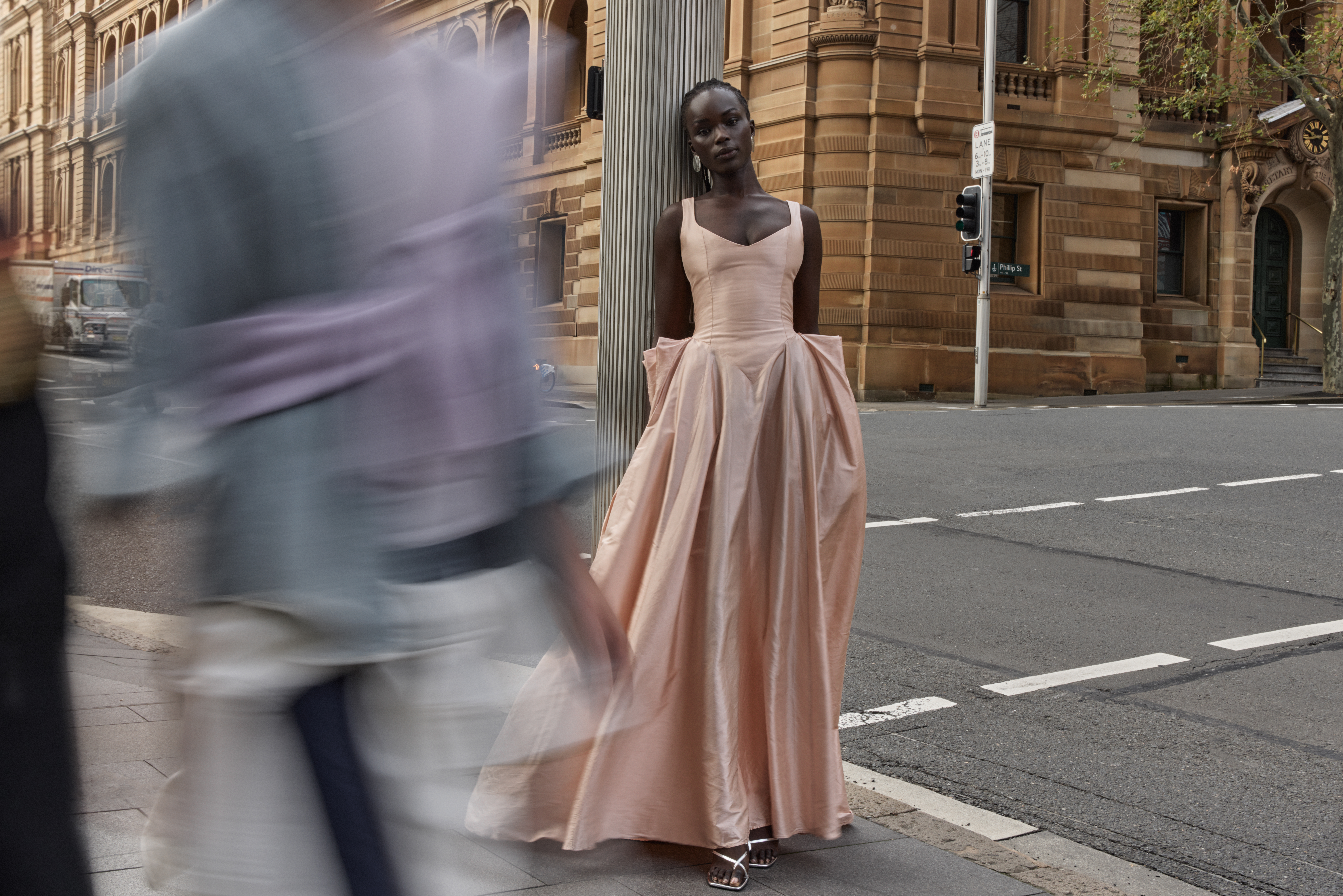 Elegant woman in a peach gown poses gracefully outdoors on a city street, with blurred passersby and a classic building facade in the background.