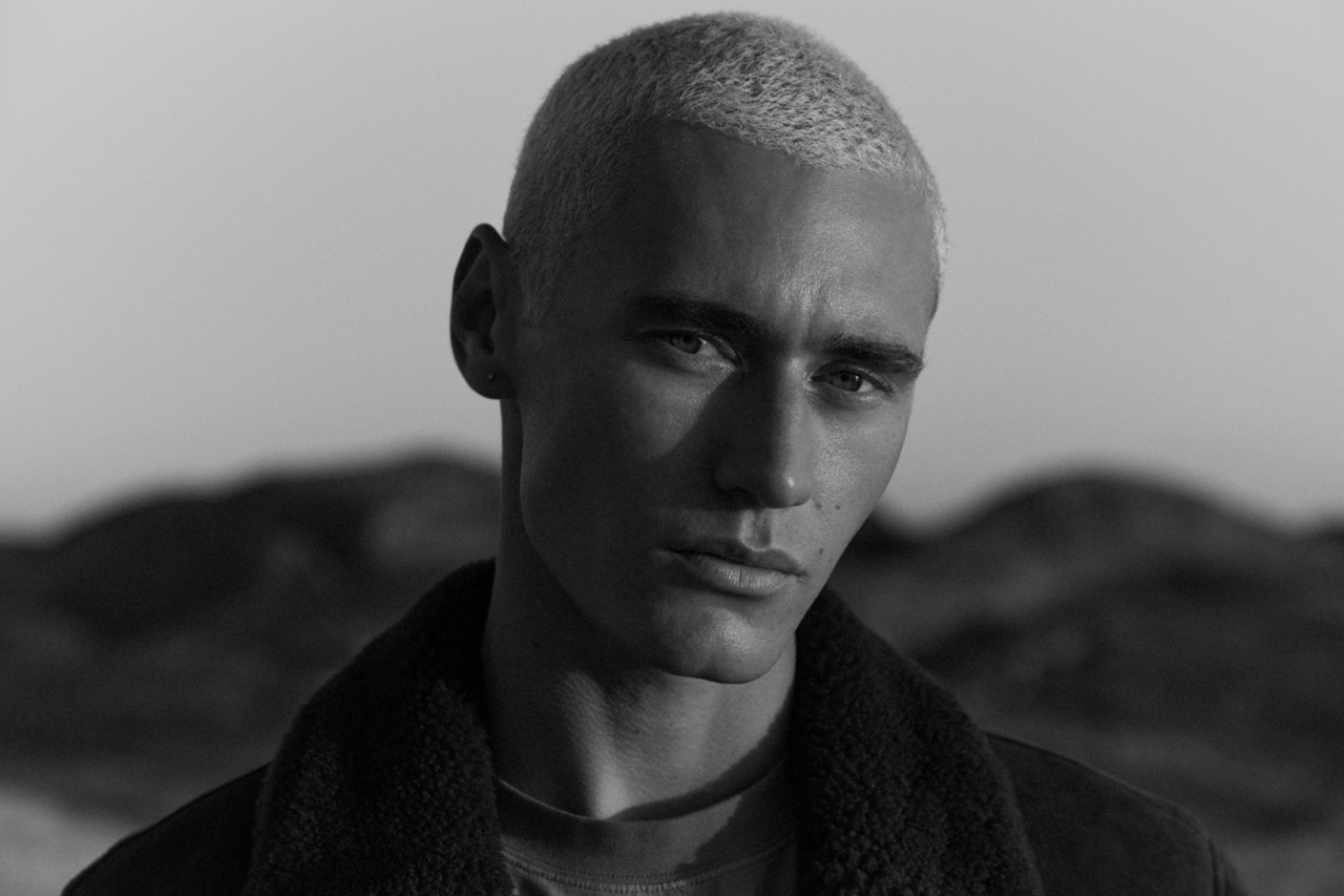 Black and white portrait of a fair-skinned adult male with a buzzcut, wearing a jacket, gazing directly at the camera against a soft outdoor background.