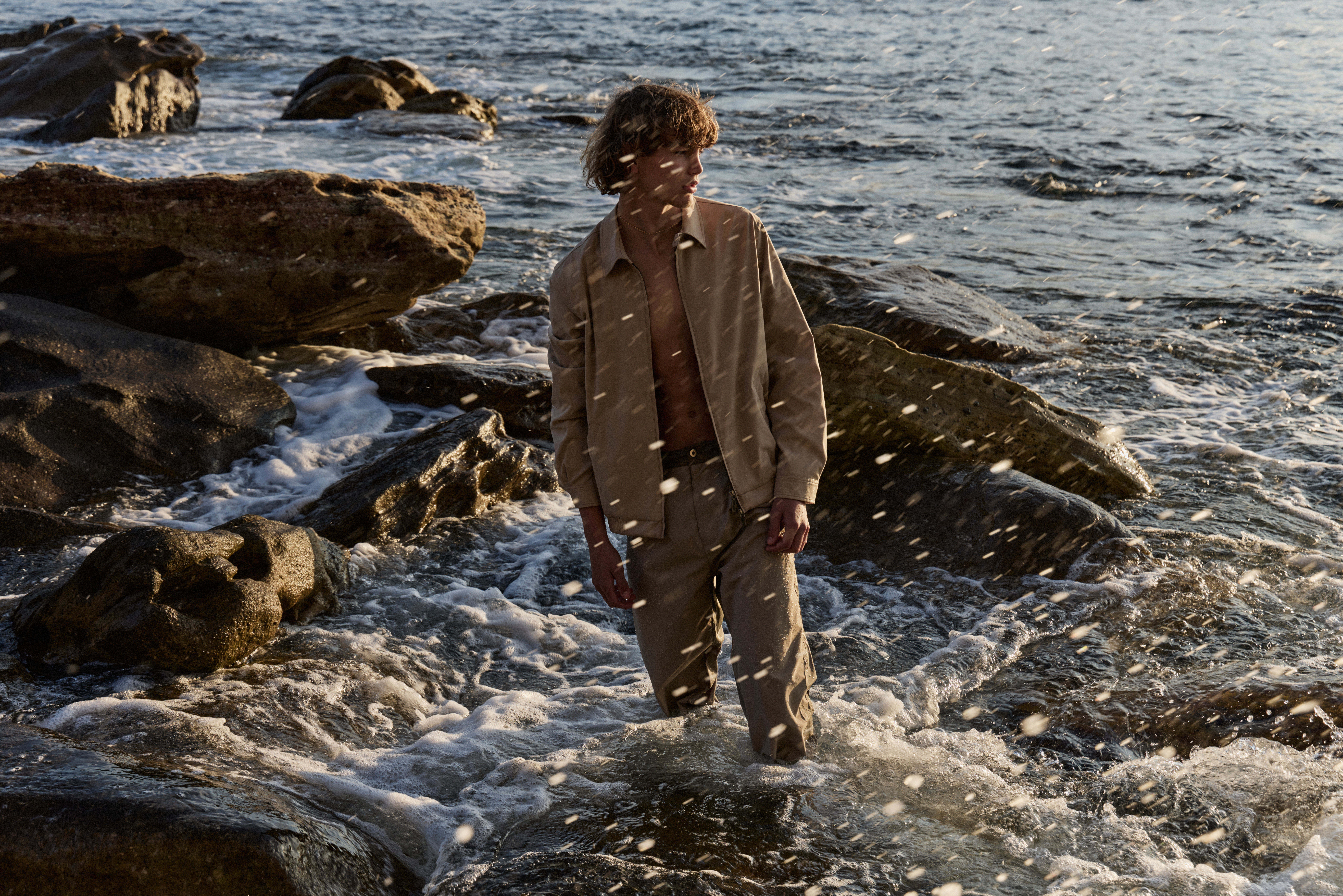 A fair-skinned man in tan clothing is walking in the ocean with crashing waves around rocky outcroppings during golden hour light.