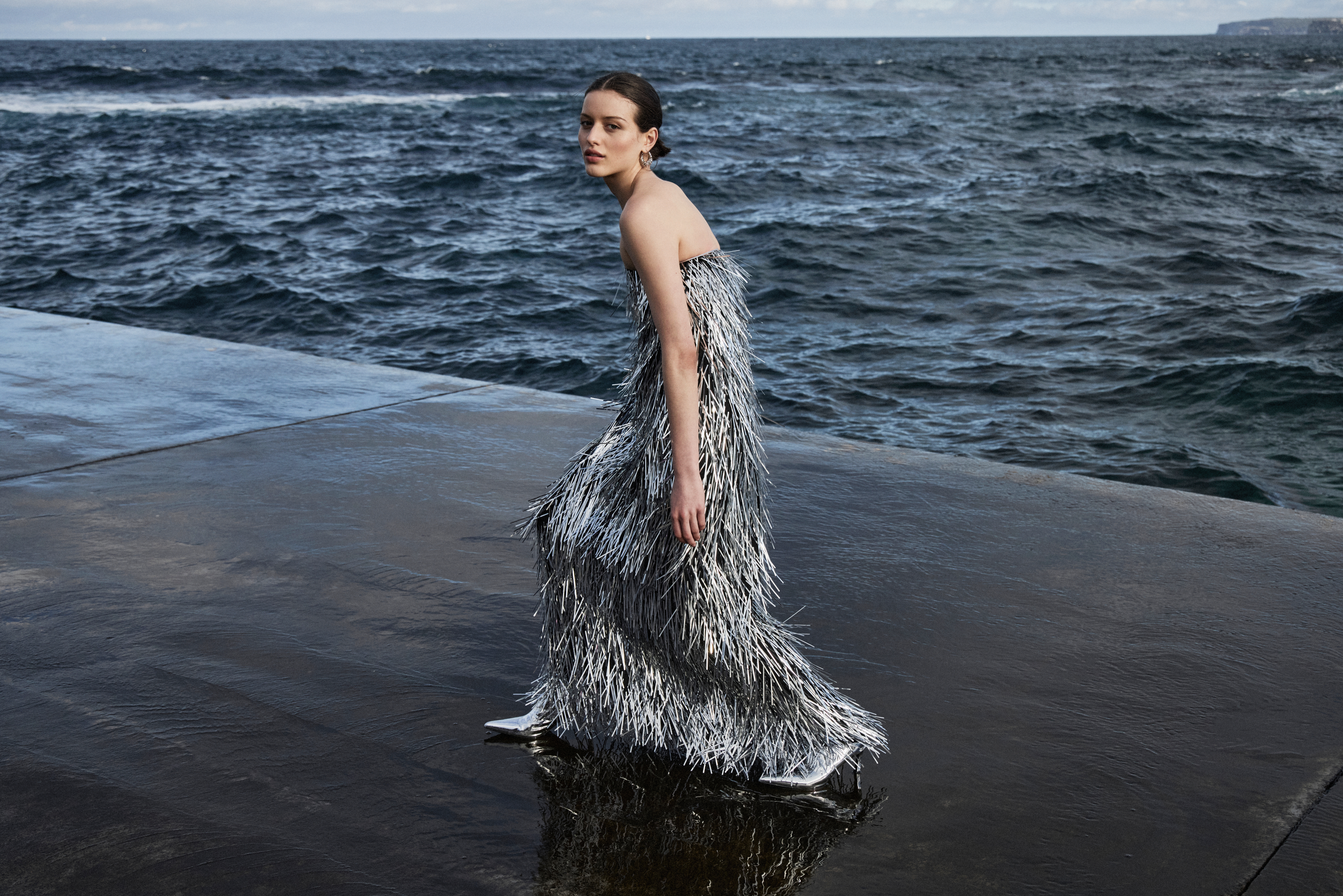 A woman in a silver fringe dress stands on a wet concrete surface with a view of the ocean; fashion editorial photography.