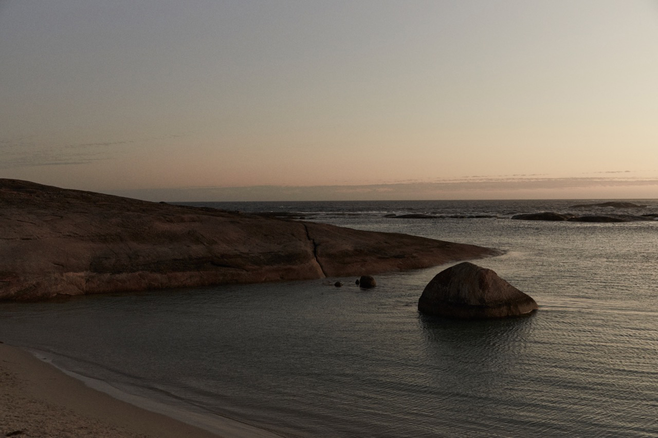 Tranquil coastal scene featuring still water and large rocks in a natural setting, under a serene sky at either sunrise or sunset.