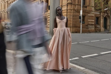 Black model in a blush pink gown leaning against a pole on a city street with motion blur in the foreground.