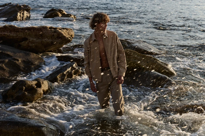 Young man standing ankle-deep in ocean surf among coastal rocks wearing a beige jacket and trousers