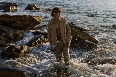 Young man standing ankle-deep in ocean surf among coastal rocks wearing a beige jacket and trousers