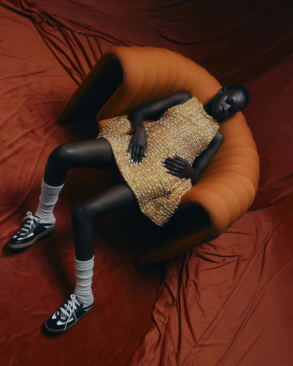 An adult female model rests in an orange chair, wearing a gold mini-dress, white socks, and black sneakers on a textured orange fabric background.