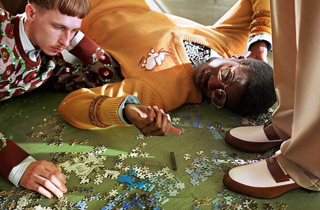 Two young men work on a jigsaw puzzle, one lying down and the other crouching on the floor, in a colorful editorial fashion shot.