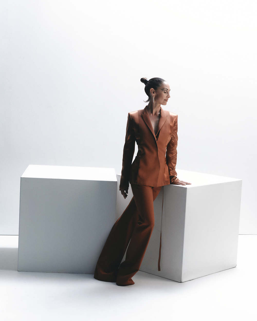 A stylish woman in a bronze pantsuit leans against white blocks in a bright studio, looking away with a sophisticated and elegant pose.