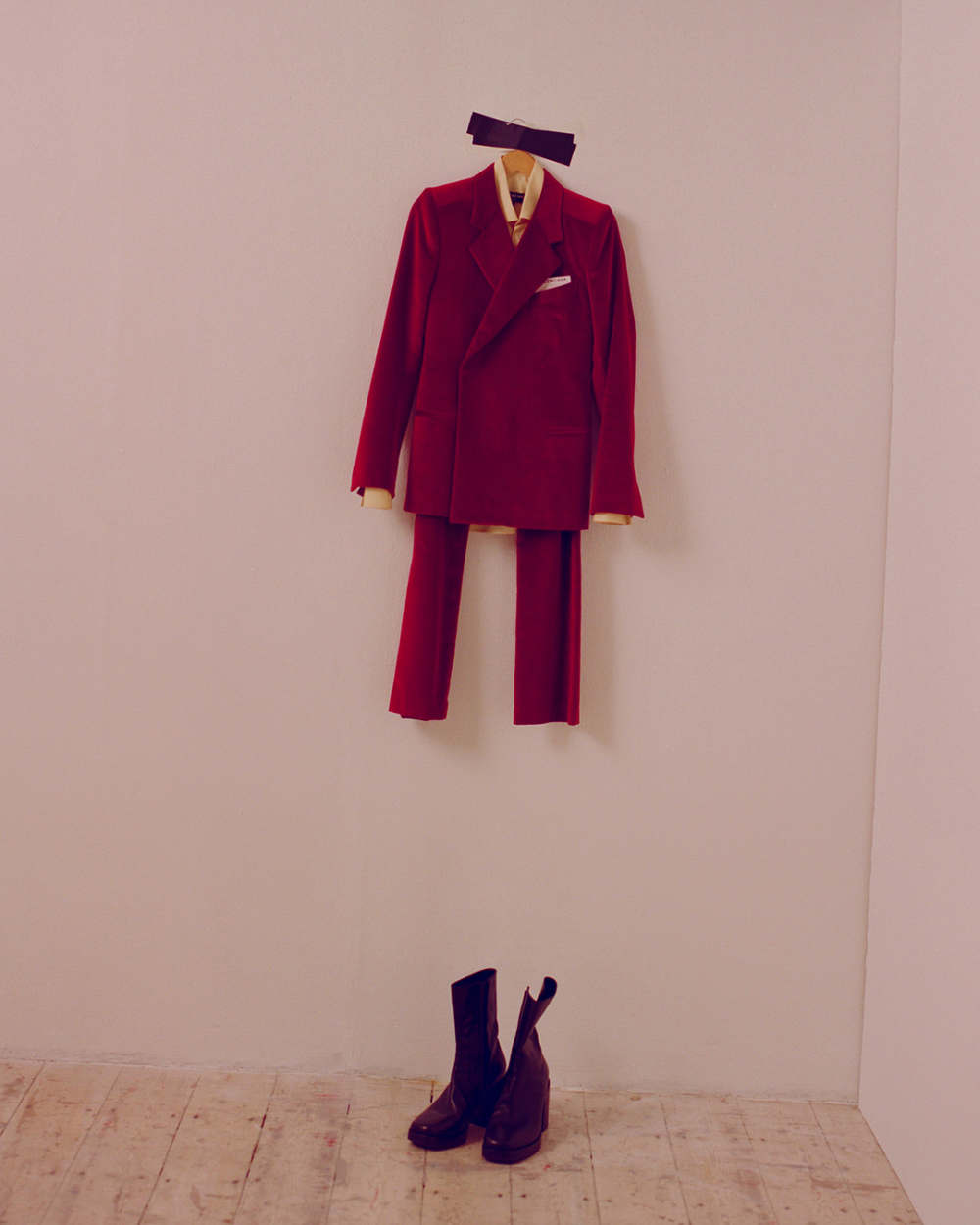 A red suit hanging against a pale pink wall with a pair of black boots on the floor, creating a fashion-forward and visually intriguing image.
