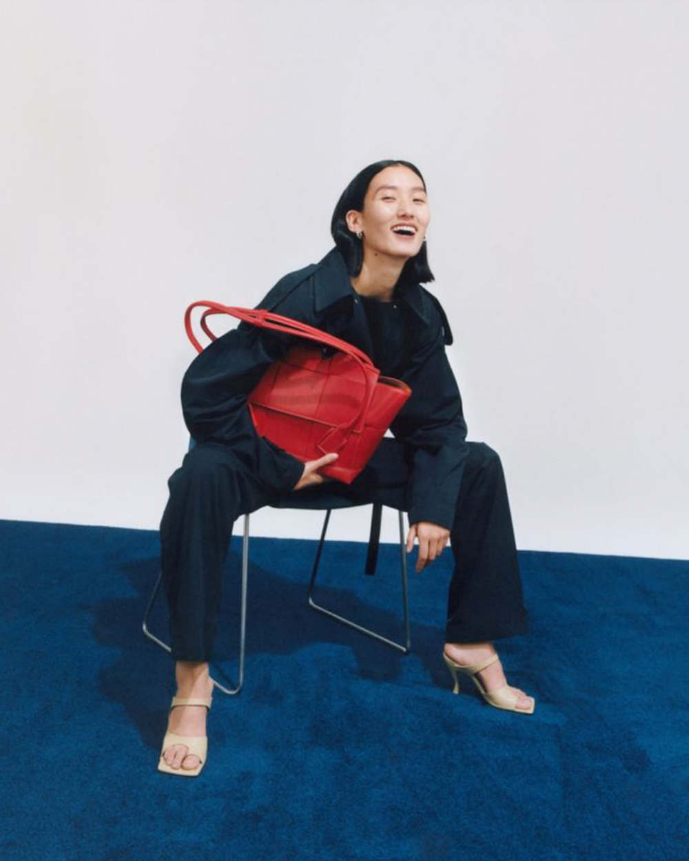 Smiling woman in a black trench coat sits on a chair and holds a red handbag in a studio with a blue carpet and a white background.