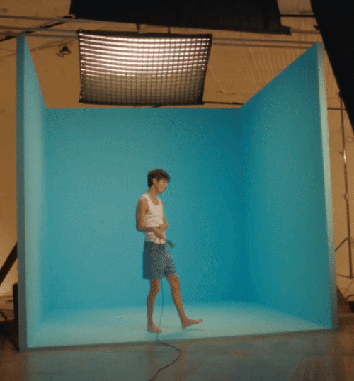 Young adult male in a tank top and shorts stands in a blue box set in a studio, holding a jump rope, lit by studio lights.