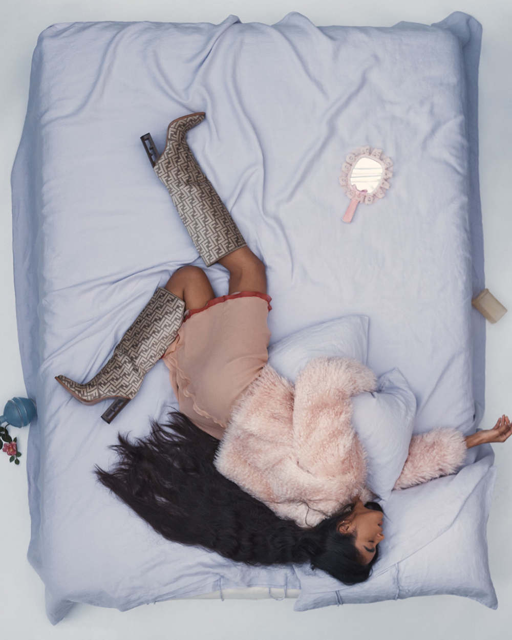 Overhead studio shot: woman lying on bed in dress and boots, surrounded by pillows and accessories, capturing a blend of fashion and comfort.