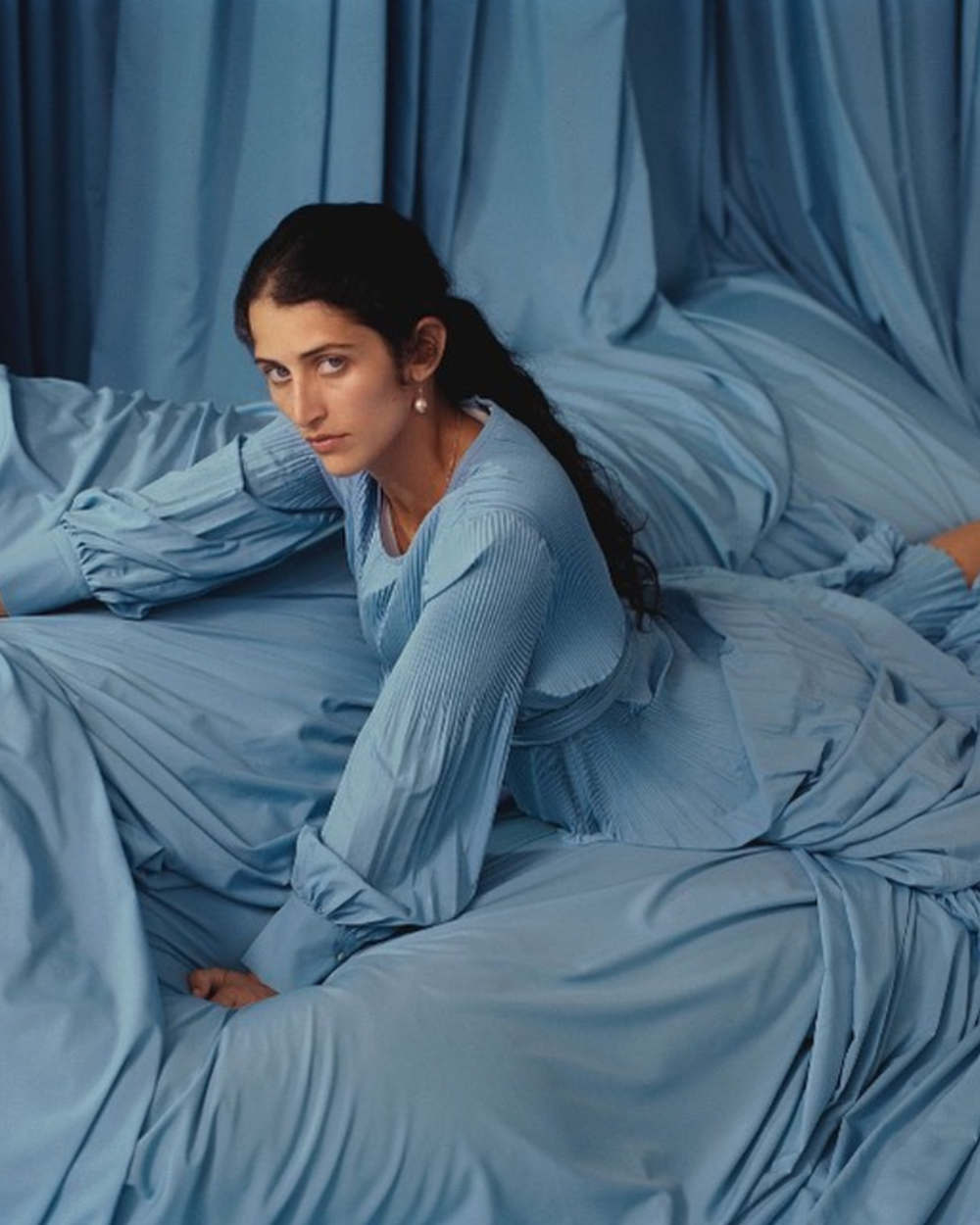 Woman with dark hair lying down on a light blue fabric, wearing a light blue matching dress, looking at the camera.