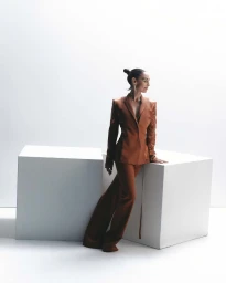 A woman in a rust-brown satin pantsuit posing between two white geometric blocks in a studio setting.
