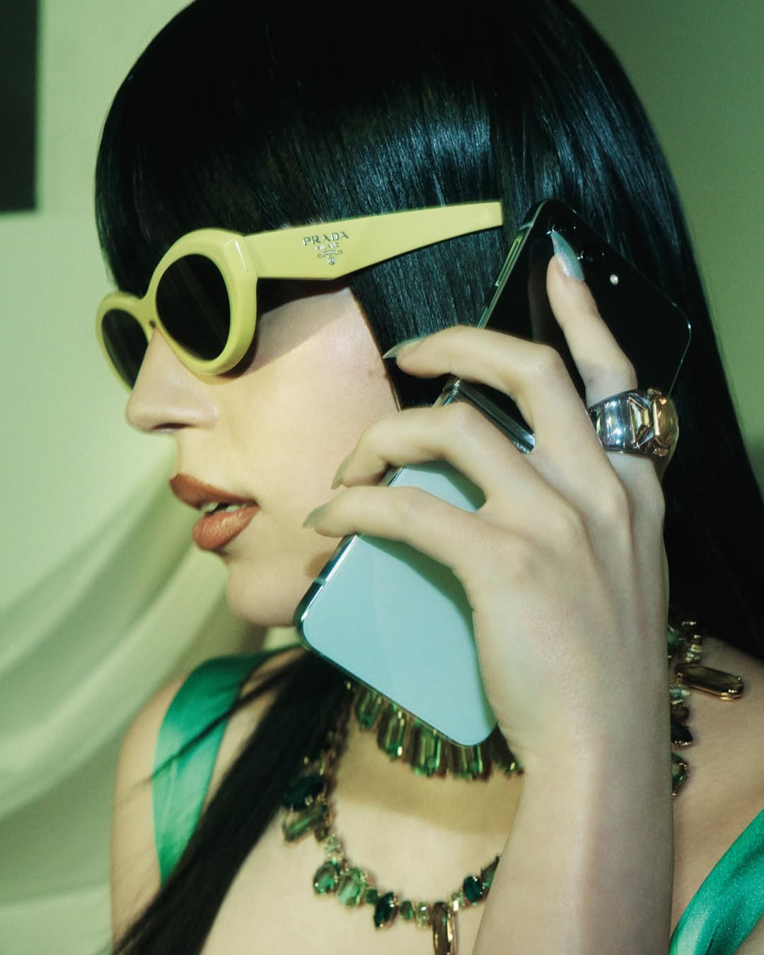Woman with black hair wearing yellow Prada sunglasses on a phone call, adorned with a chunky jewel necklace and statement rings.