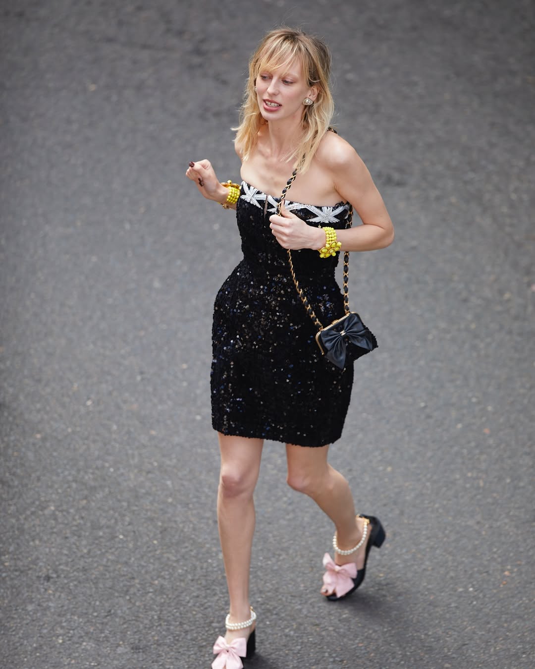 Woman in a black sequin dress with yellow and white accents and pink bow heels walking on a grey surface. She is carrying a purse.