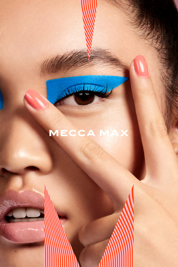 Asian adult female close-up with bright blue eye makeup and coral nail polish holds fingers up in front of eye. Mecca Max text present.