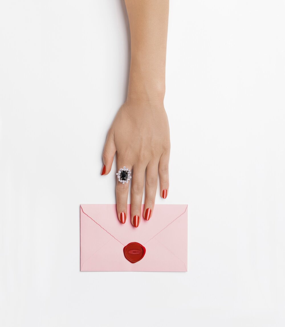 Woman's hand with a ring reaching for a pink envelope with a red wax seal on a white background in a studio shot.
