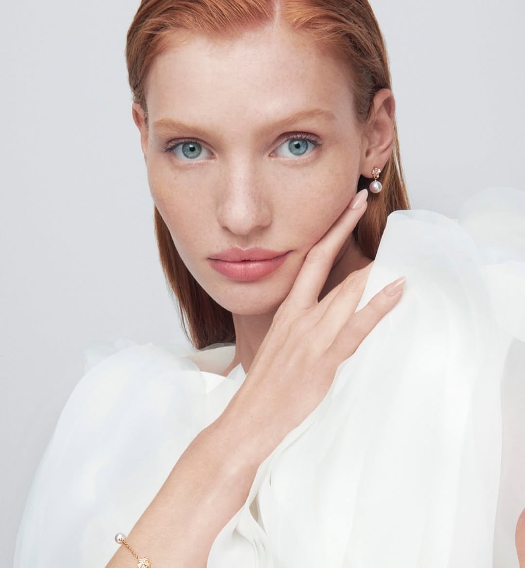 Close-up of a fair-skinned, red-haired woman wearing pearl earrings and a bracelet. She is wearing a white blouse against a light background.