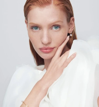 Close-up of a fair-skinned, red-haired woman wearing pearl earrings and a bracelet. She is wearing a white blouse against a light background.