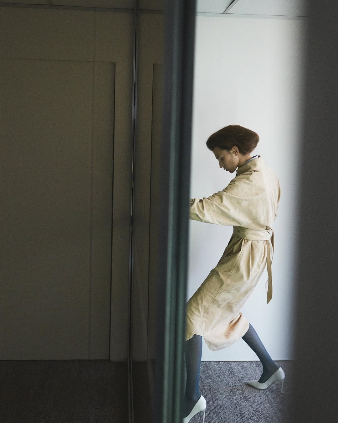 A woman in a trench coat and heels pushes a glass door in a high-key fashion shot, minimalist aesthetic, neutral colors, and contemporary design.