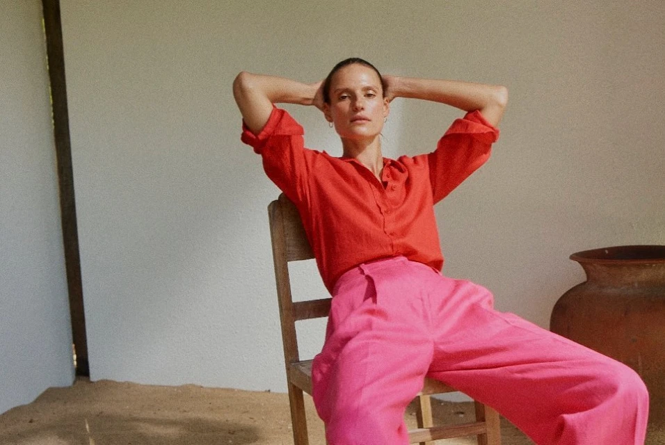 Adult woman in casual fashion, posing while seated in a minimalist indoor environment, showcasing red top and pink pants.