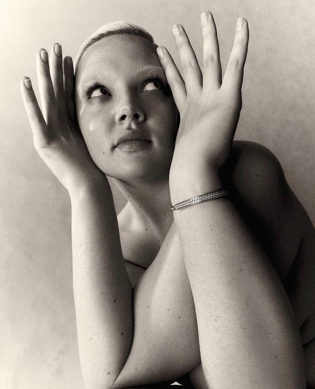 Monochrome portrait of a woman with blonde hair and a bracelet posing with her hands near her face in a photo studio.