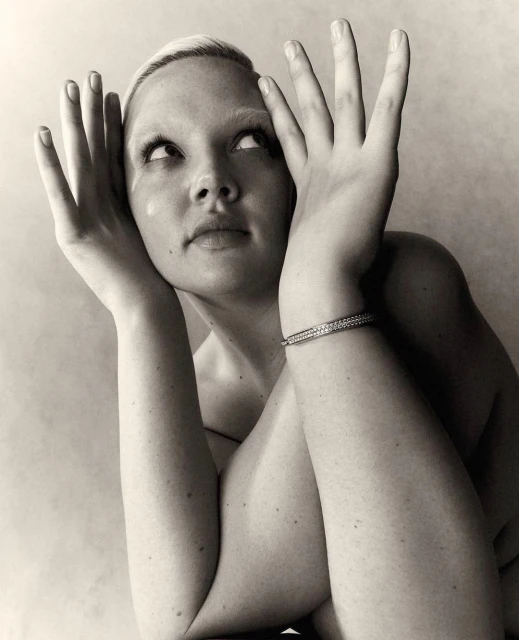 Monochrome portrait of a woman with blonde hair and a bracelet posing with her hands near her face in a photo studio.