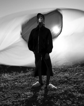 Monochrome fashion shot of a woman in a jacket and skirt, standing in a grassy field with a blurred white shape in the background.