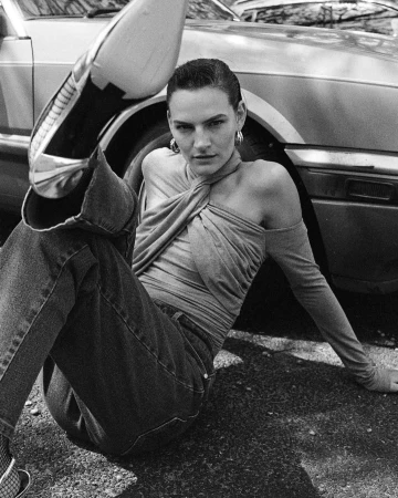 Black and white portrait of a woman seated on the ground with one leg raised towards the camera, leaning against a parked car.