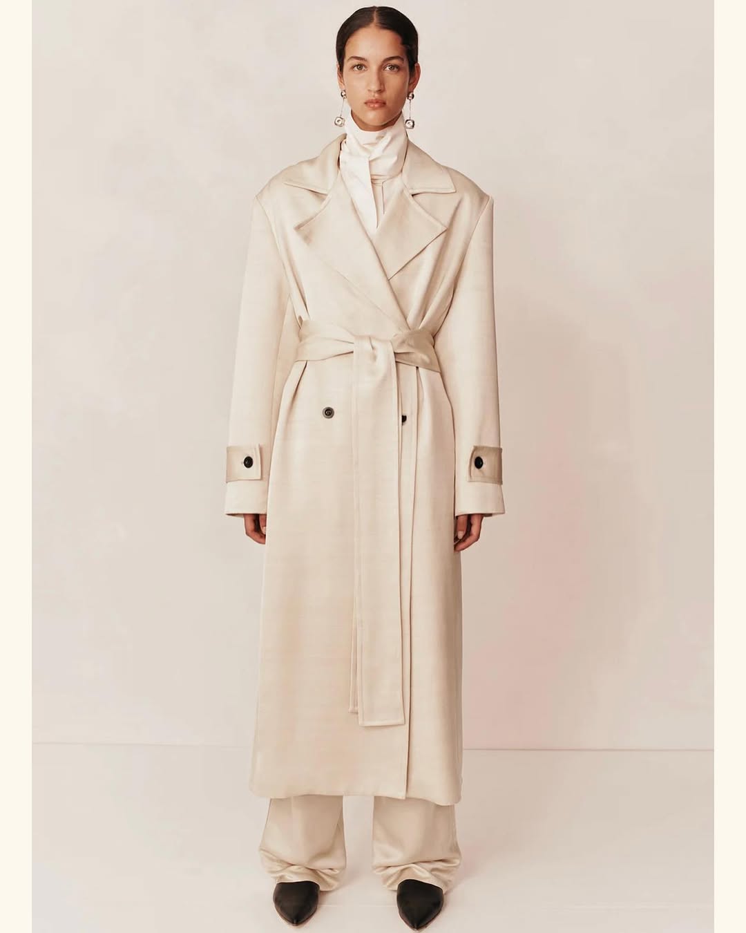 A woman in a cream-colored trench coat and wide-legged trousers poses in a studio against a light background for a fashion editorial.
