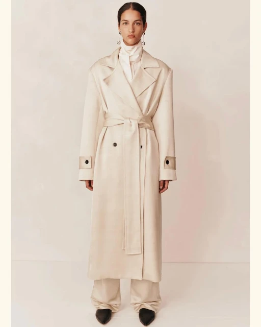 A woman in a cream-colored trench coat and wide-legged trousers poses in a studio against a light background for a fashion editorial.