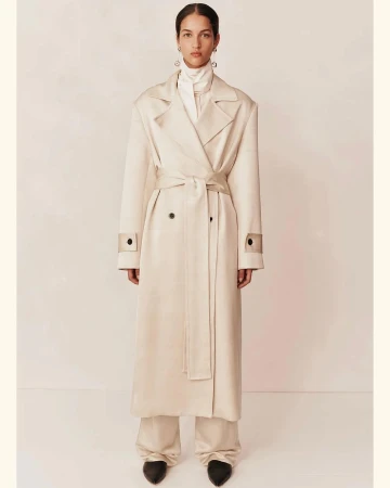 A woman in a cream-colored trench coat and wide-legged trousers poses in a studio against a light background for a fashion editorial.