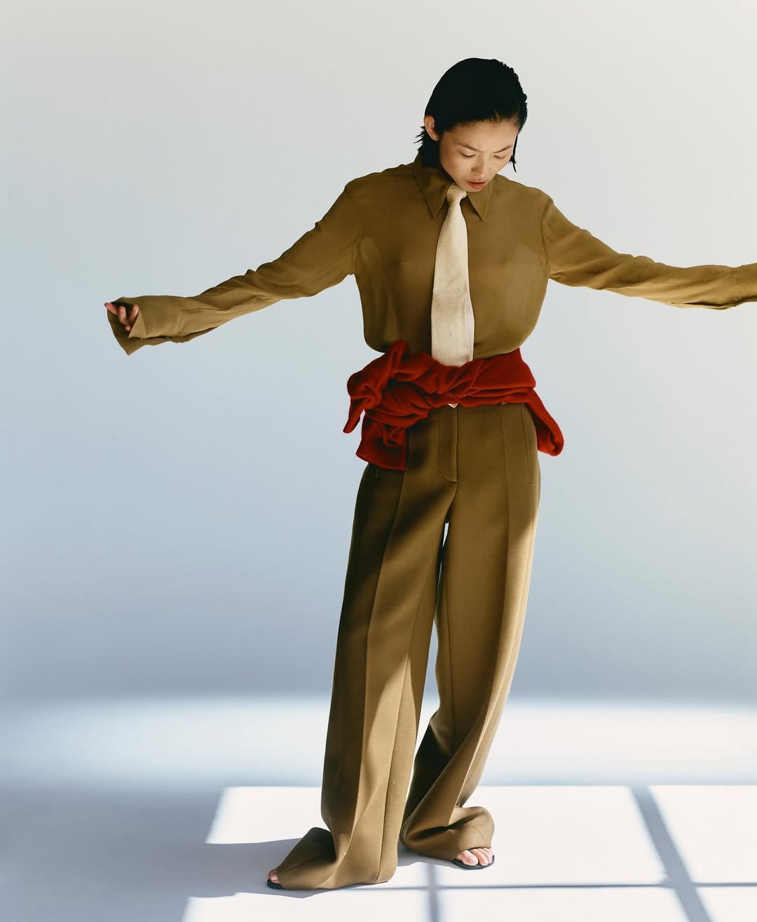 Adult female fashion model poses in olive button-down shirt, light tie, brown trousers, and red sweater against a neutral backdrop.