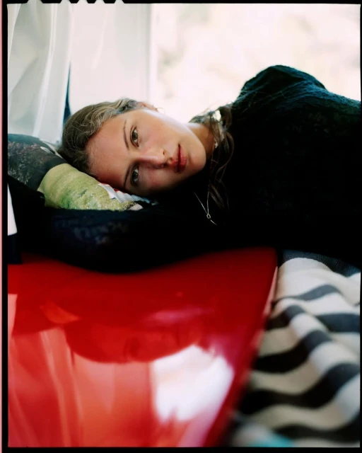 Portrait of a young woman lying on a red surface with striped fabric, looking at the camera with a neutral expression in a domestic setting.