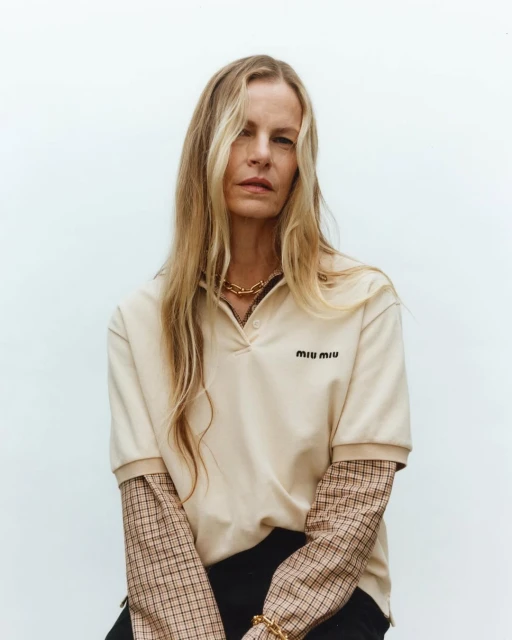 Portrait of an adult female with long blonde hair wearing a beige polo shirt with black pants against a plain white background.