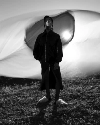 Black and white portrait of a model in a dark zip jacket and skirt standing outdoors against a sweeping fabric backdrop.
