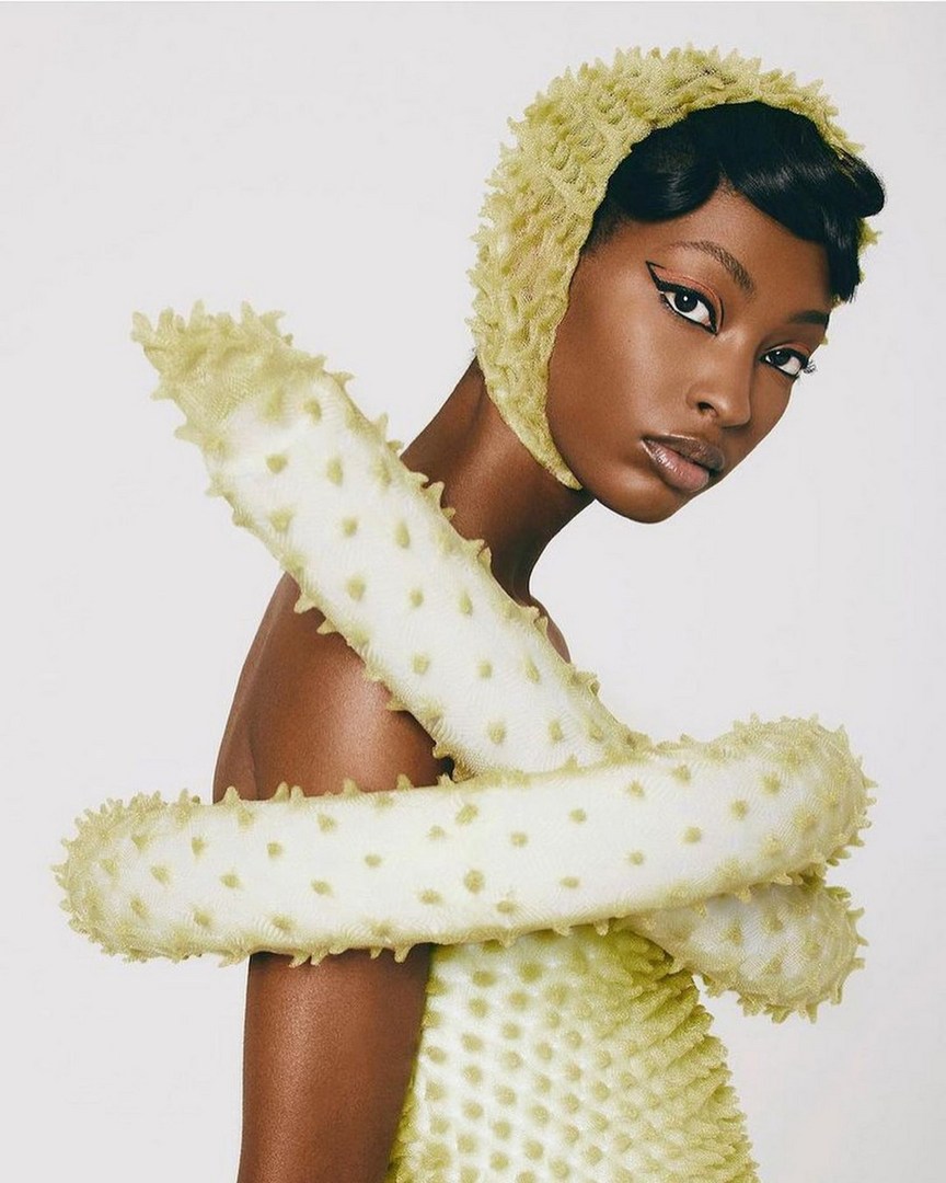 Dark-skinned woman in studio wearing textured, pale yellow dress with matching hood and tubular, tentacled forms around her arms.
