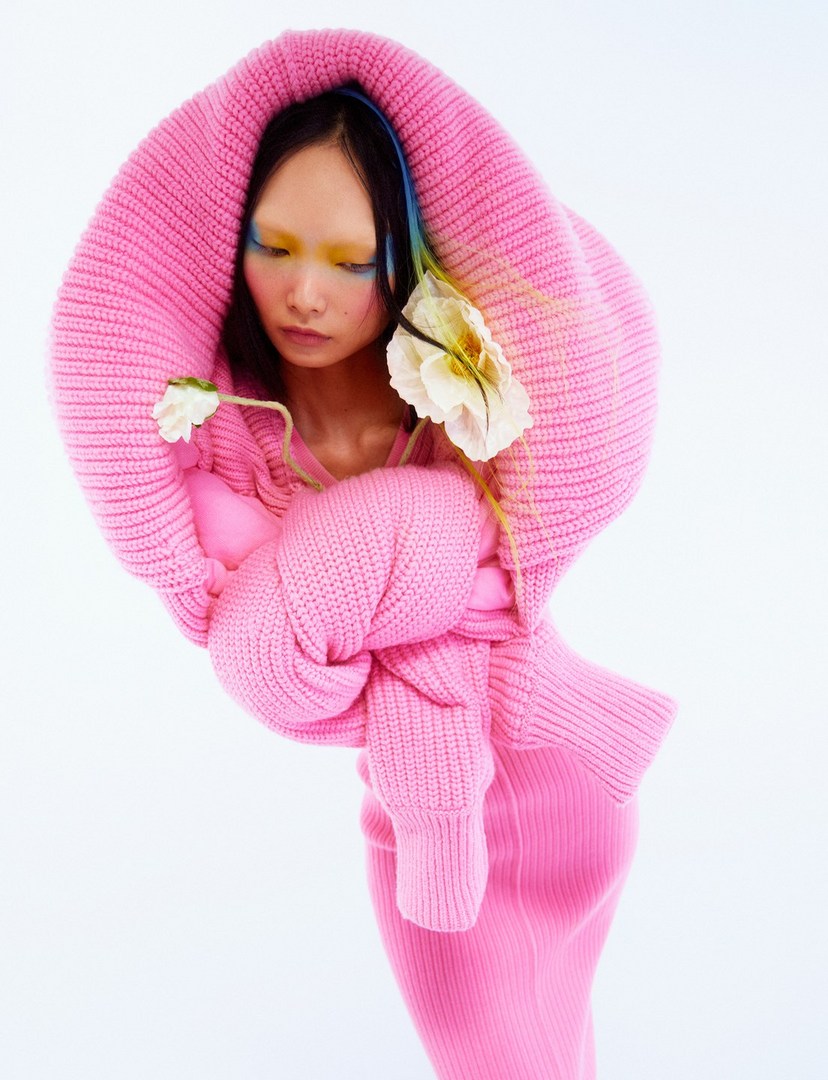 Woman in pink hooded knitwear with white flowers in a studio setting. Fashion-forward editorial image with a clean white background.