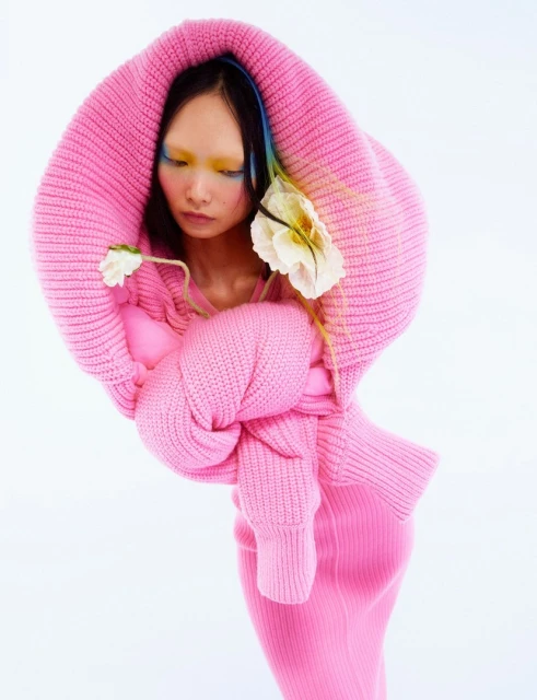 Woman in pink hooded knitwear with white flowers in a studio setting. Fashion-forward editorial image with a clean white background.