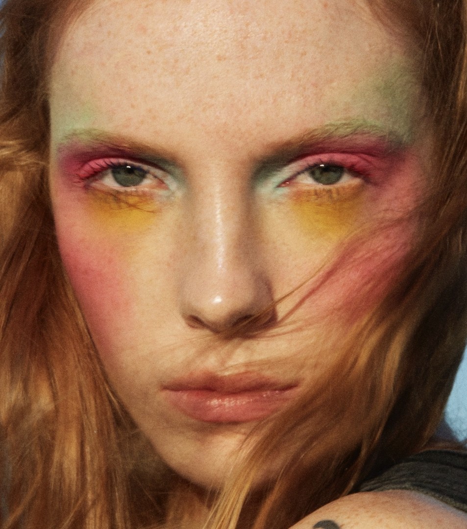 Close-up of a woman's face with vibrant rainbow makeup, including green, pink, and yellow eyeshadow, highlighted cheekbones, and red hair.