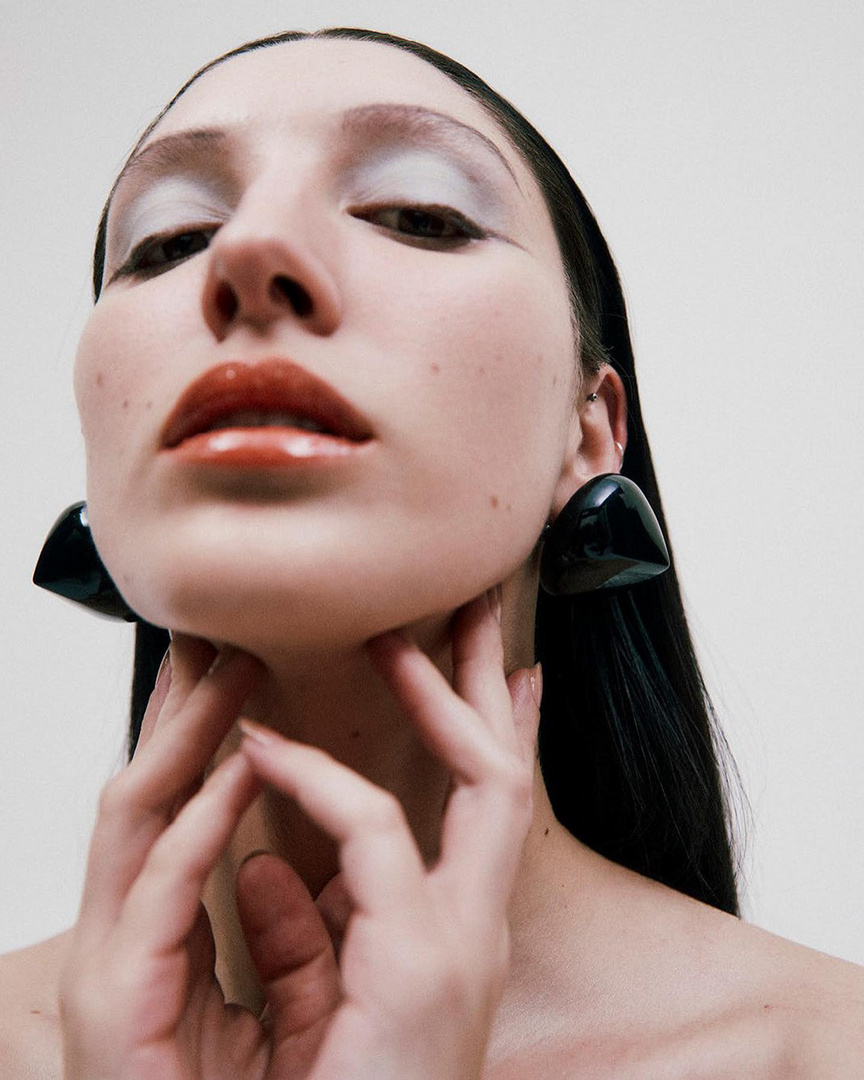 Close-up studio shot of a woman with glossy lips and large black triangle earrings touching her neck with both hands.