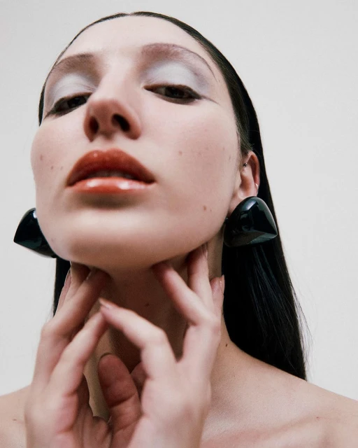 Close-up studio shot of a woman with glossy lips and large black triangle earrings touching her neck with both hands.