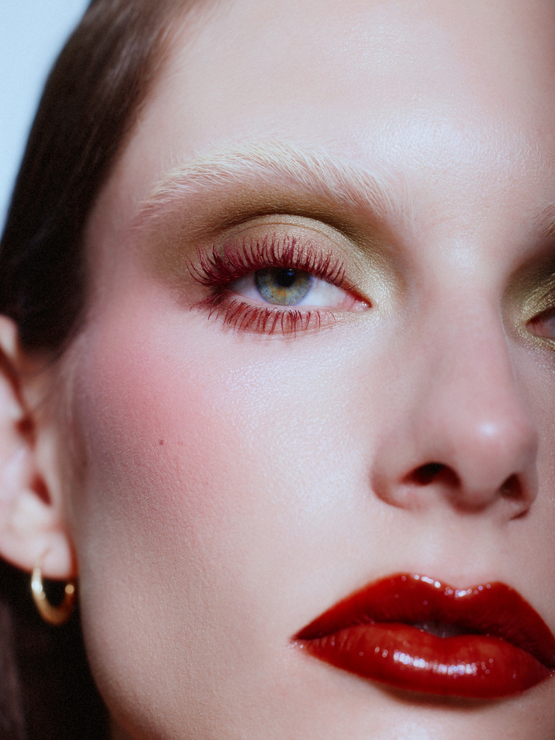 Close-up portrait of a woman with light skin, red lips and mascara, and green eyeshadow, set against a soft blue background.