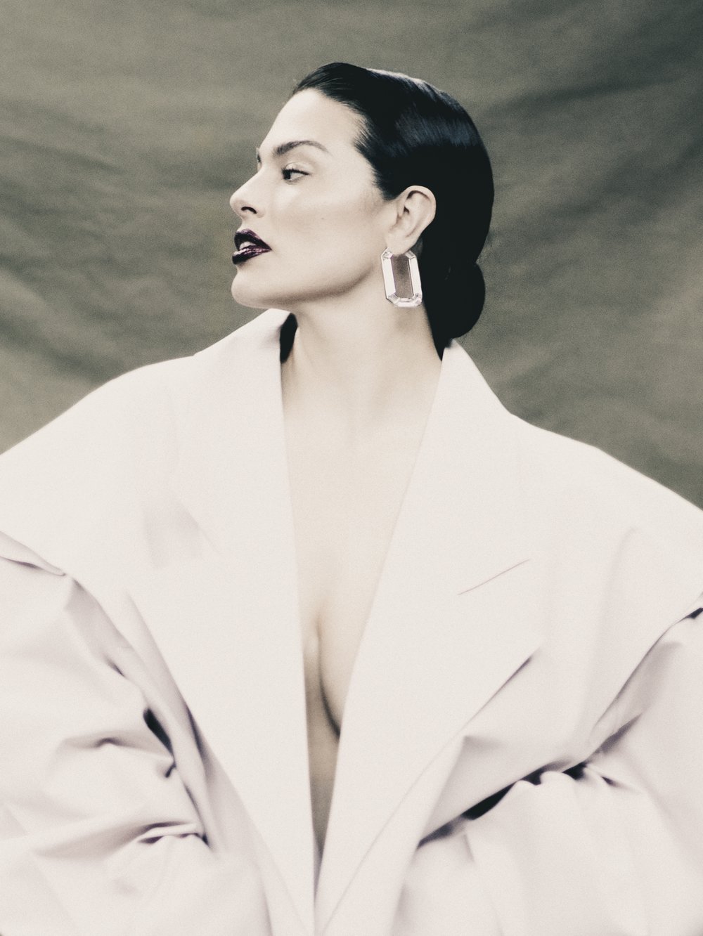 Woman in profile wearing a white jacket and earrings against a draped background in a studio portrait showcasing fashion and beauty.