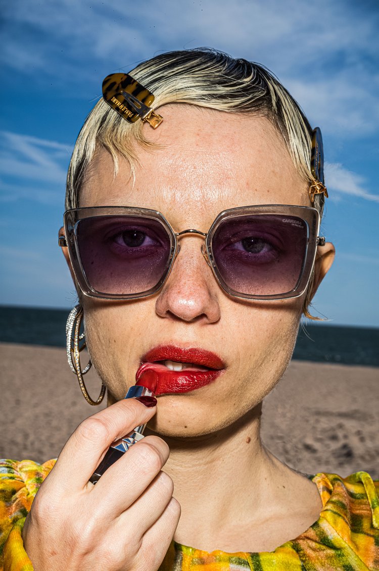 Woman applying red lipstick in a close-up shot on a sandy beach with a blue sky, wearing sunglasses and hair clips.