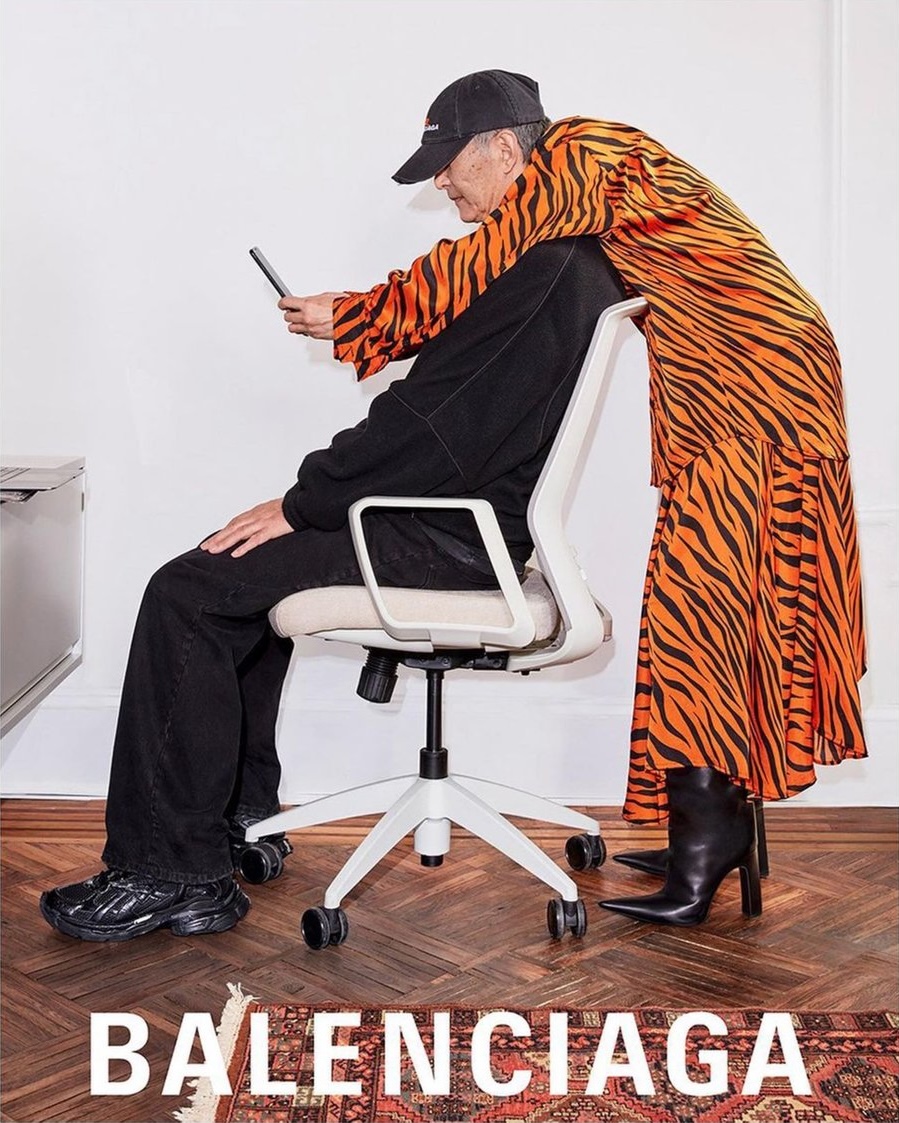 Fashion editorial featuring an adult female in an orange tiger-striped dress and an adult male in a black tracksuit in a studio.