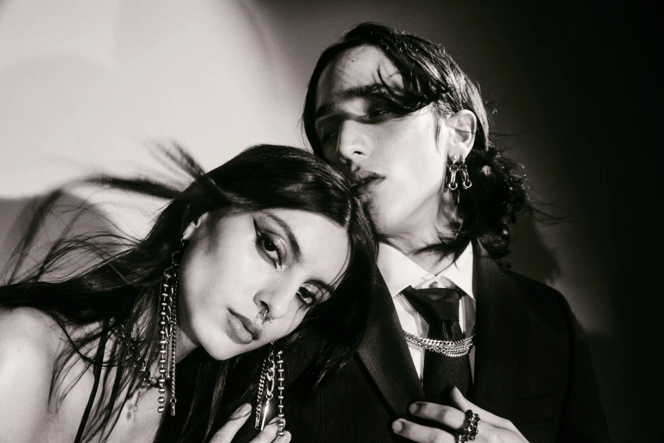 Monochrome portrait of a fashionable couple, a woman with dramatic eye makeup and a man in a suit, posing in a studio setting.